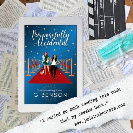 On an iPad, the cover of Purposefully Accidental by G Benson on a backdrop of book pages on a wooden floor. On the upper right side, next to the iPad, are a clapboard, a fidget ring, and a surgeon's mask. At the bottom of the image, slightly covering the iPad, a strip of torn paper on this is a quote: "I smiled so much reading this book that my cheeks hurt." and a url: www.judeinthestars.com