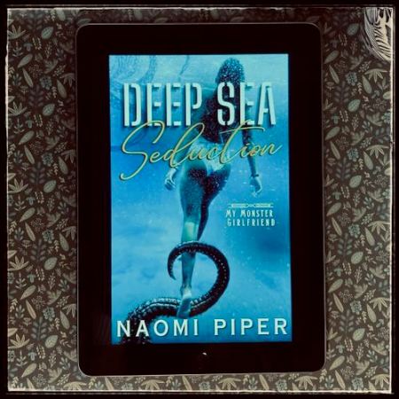 On a darkish flowery background, an iPad with the cover of Deep Sea Seduction by Naomi Piper.