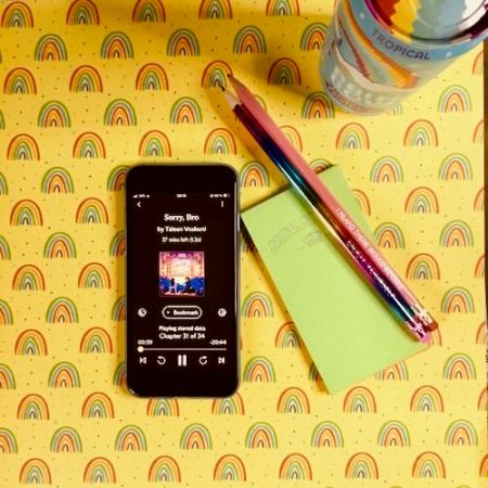 An iPhone with the cover of Sorry, Bro by Taleen Voskuni, narrated by Christine Mirzayan, on a yellow background with small rainbows. Next to the phone is a can of rainbow soda, a small green notebook, and two pencils, one with the words "Enjoy yourself" and one with "I heard there would be cake".