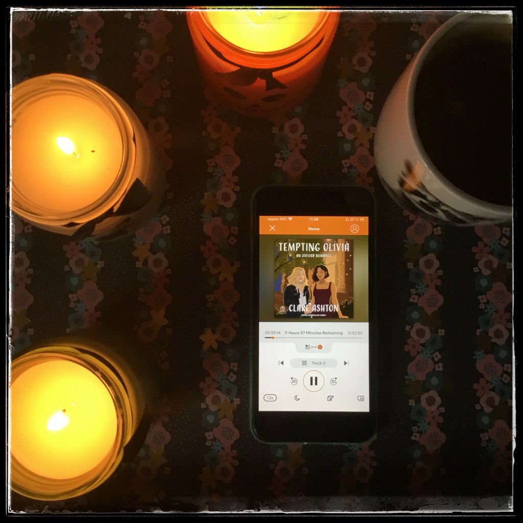 On a dark flowery background, an iPhone with the cover of Tempting Olivia (Oxford Romance #2) by Clare Ashton, narrated by Shiloh Grey, surrounded by three lit candles and a mug of tea with the picture of a raccoon.