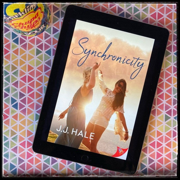 On a background of multicoloured triangles, an iPad with the cover of Synchronicity by J.J. Hale, next to a can of soda.
