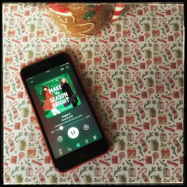 On a festive Christmas background, an iPhone with the cover of Make the Season Bright by Ashley Herring Blake, narrated by Kristen DiMercurio, and next to it, a gingerbread man mug.