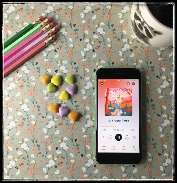 An iphone with the cover of The View from the Top by Rachel Lacey, narrated by Quinn Riley on a flowery background. Next to the phone are a few pencils, multicoloured heart candies, and a mug of tea.