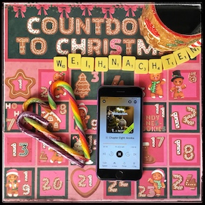An iPhone with the cover of Merry Weihnachten by E. J. Noyes, narrated by Abby Craden, next to a couple of rainbow candy canes and a gingerbread man mug. The background is a pinkish countdown to Christmas, with various drawings, and below the word "Christmas", Scrabble letters spelling "Weihnachten".