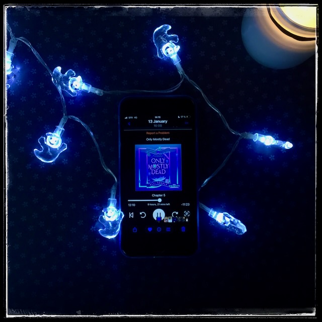 An iPhone with the cover of Only Mostly Dead (Afterlife Incorporated #1) by Alli Temple, narrated by Emily Lawrence, on.a dark background. A string of ghost fairy lights surrounds the phone and a lit candle occupies the upper right corner.
