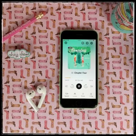 On a pink background with a cowgirl boots pattern, an iPhone playing Graceless by Ruby Landers, narrated by Quinn Riley. Next to it are a pink pencil with the words "I need more middle fingers, a they/them button, a pair of AirPods, and a can of soda.