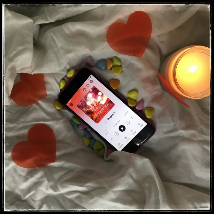 On white creased sheets, an iPhone with the audiobook of The Perfect Matchmaking by Milena McKay, narrated by Abby Craden, surrounded with colourful heart-shaped candy and red paper hearts. On the right side, a white candle is shining.