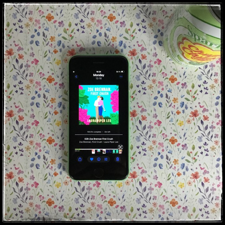 On a flowery background, an iPhone with the cover of Zoe Brennan, First Crush by Laura Piper Lee, narrated by Lindsey Dorcus. There's also a green soda can in the top right corner.