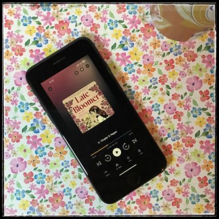 On a bright flowery background, an iPhone with the cover of Late Bloomer by Mazey Eddings, narrated by Ellie Gossage.