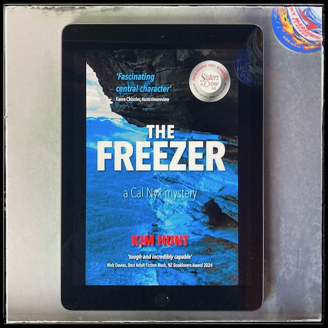 On a greyish background, an iPad with the cover of The Freezer by Kim Hunt. In the top right corner, a glimpse of a colorful can of soda.