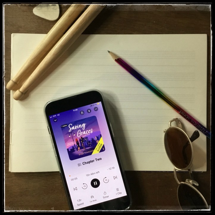 On a wooden background, an iPhone with the cover of Saving Graces by Ruby Landers, narrated by Quinn Riley, on music paper, next to drumsticks, a guitar pick, a rainbow pencil and sunglasses.